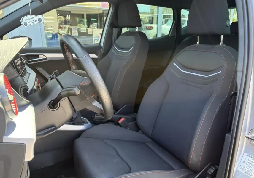 Vue intérieure côté conducteur du SEAT Arona 2024, sièges textile noirs avec surpiqûres rouges et volant sport cuir.
