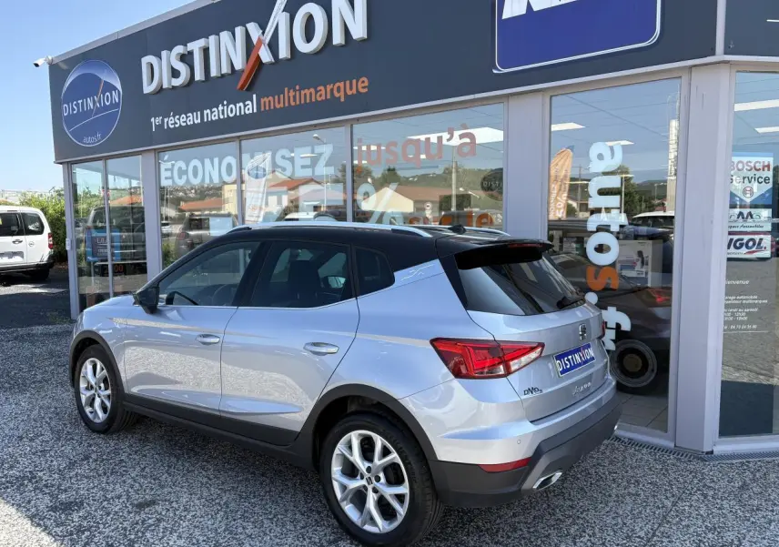 Vue 3/4 arrière droite d’un SEAT Arona 2024 gris clair avec jantes alliage et vitres teintées, devant un showroom.