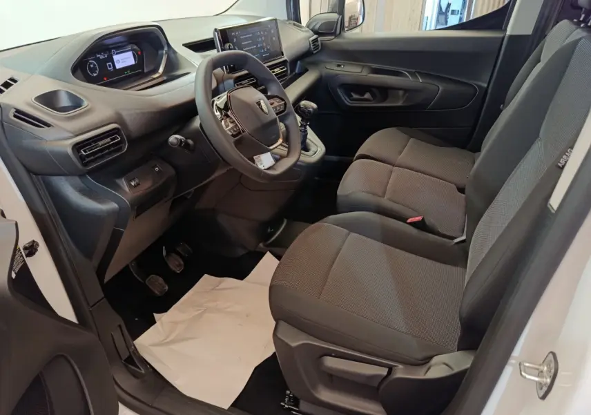 Vue intérieure côté conducteur du Peugeot Partner blanc 2025, montrant le tableau de bord et les sièges tissu gris.