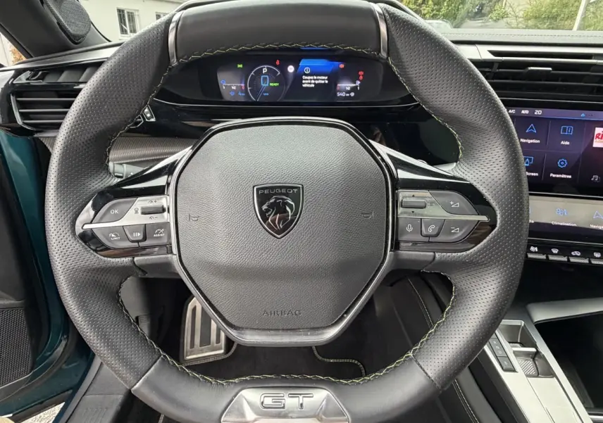 Vue rapprochée du volant cuir noir GT avec logo Peugeot et tableau de bord numérique du Peugeot 408 PHEV 225 bleu.
