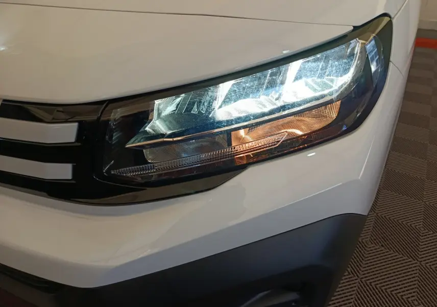 Gros plan sur le phare avant droit du Peugeot Partner blanc 2025 avec calandre noire et détails lumineux LED.