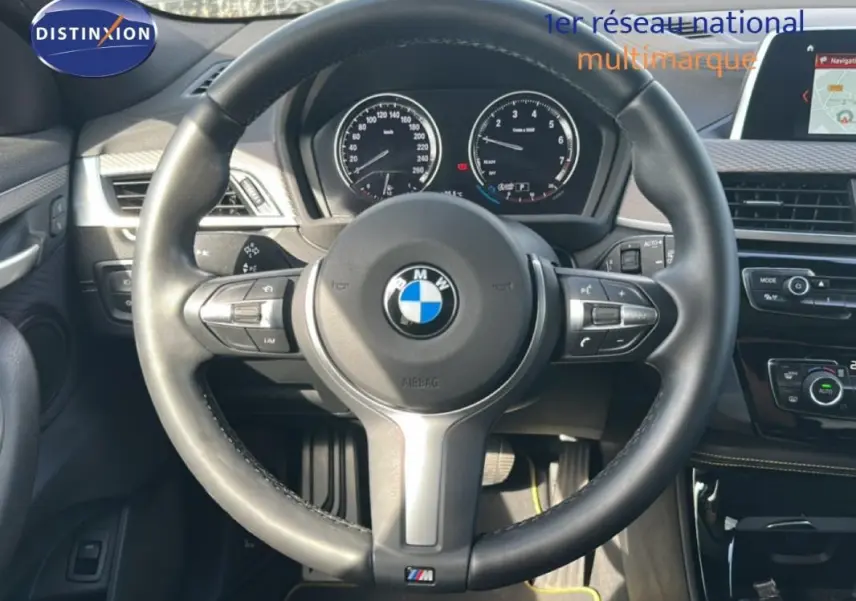Vue centrée sur le volant sport BMW d’un X2 blanc 2018 avec tableau de bord et écran GPS visibles.
