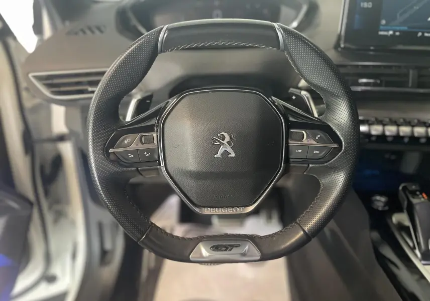 Vue rapprochée du volant cuir perforé noir avec logo GT du Peugeot 3008 blanc nacré, tableau de bord en arrière-plan.