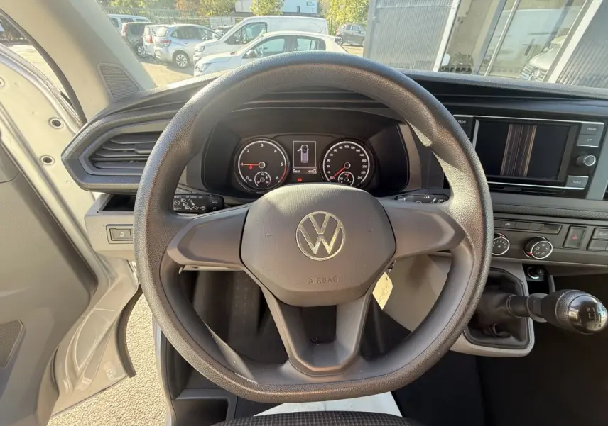 Vue intérieure centrée sur le volant noir et le tableau de bord du Volkswagen Transporter gris clair, avec levier de vitesses manuel visible.