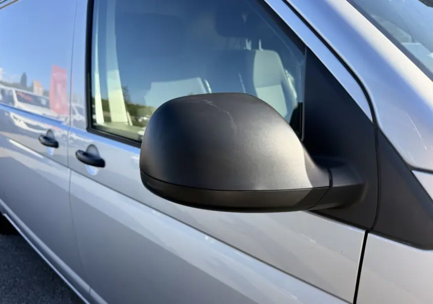 Vue rapprochée du rétroviseur côté conducteur d’un Volkswagen Transporter gris clair 2022, avec portières visibles.