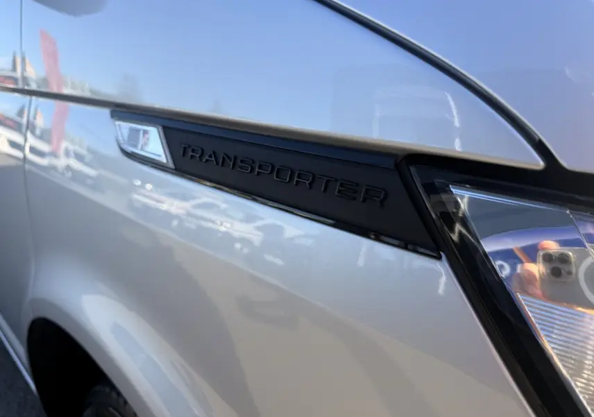 Gros plan sur l'aile avant gauche gris clair du Volkswagen Transporter avec logo noir "TRANSPORTER" et phare partiel visible.
