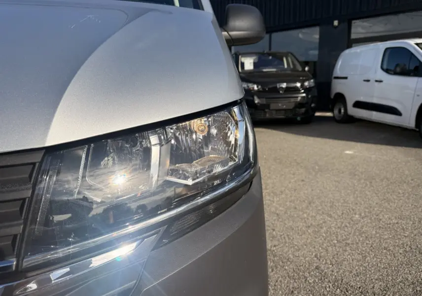 Gros plan sur le phare avant gauche du Volkswagen Transporter gris clair, avec calandre noire visible.