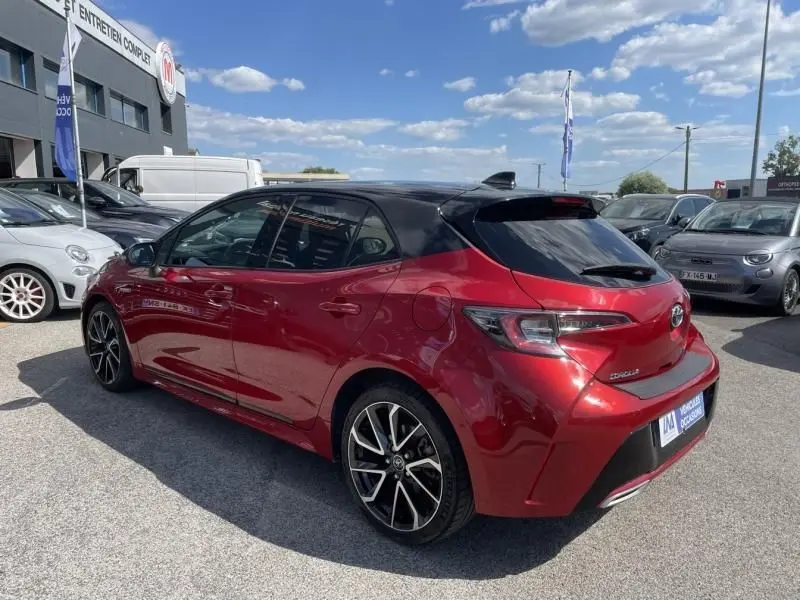 Vue 3/4 arrière droite d'une Toyota Corolla 184 H Collection rouge métallisé avec toit noir, roues alliage et feux arrière distinctifs.