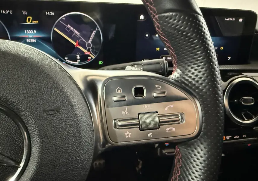 Gros plan sur le volant cuir noir avec surpiqûres rouges et commandes multifonctions d'une Mercedes Classe A gris foncé, tableau de bord digital visible.