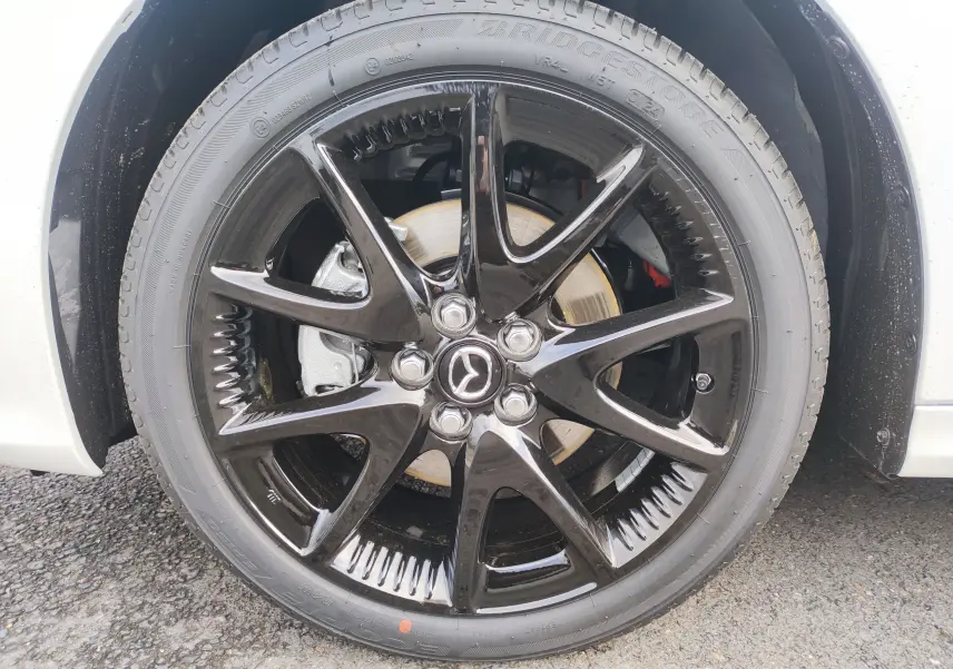 Gros plan sur la roue avant droite de la Mazda2 Hybrid 2024 blanc métallisé avec jante noire brillante et logo Mazda central.