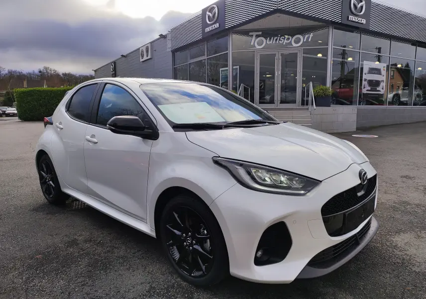 Vue 3/4 avant gauche d'une Mazda2 Hybrid blanc métallisé avec jantes noires devant un concessionnaire Mazda.