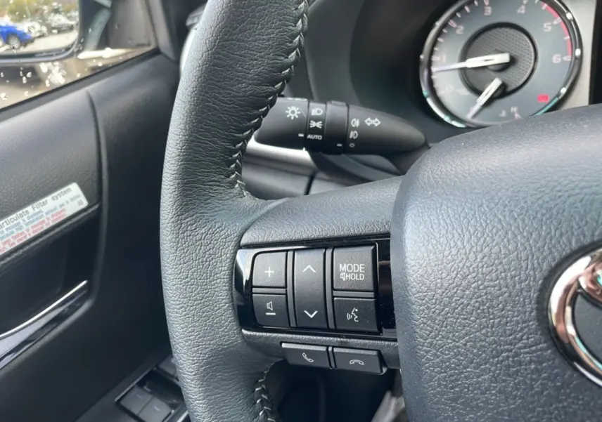 Gros plan sur les commandes au volant à gauche d'un Toyota Hilux 2025, finition Bronze Imperial, avec tableau de bord en arrière-plan.