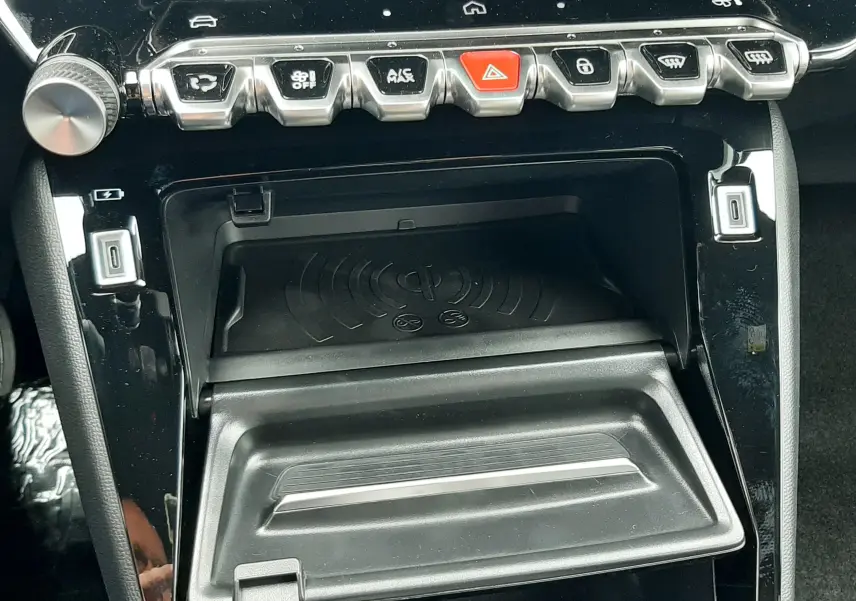 Gros plan sur la console centrale noire brillante du Peugeot 2008 1.2 Hybrid 145, avec boutons de commande et chargeur sans fil.