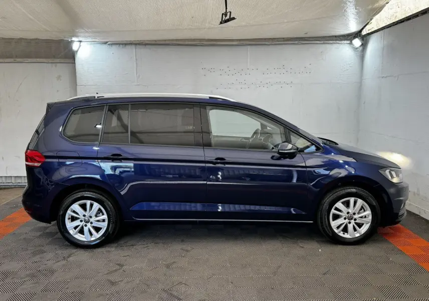Vue de profil côté gauche d'un Volkswagen Touran bleu 2024 stationné en intérieur sur sol carrelé.
