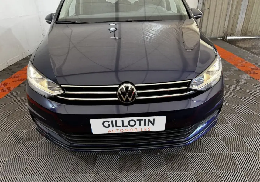 Vue avant rapprochée d’un Volkswagen Touran bleu 2024 avec calandre chromée et phares allumés dans un garage.