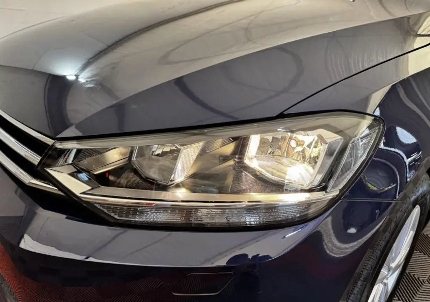 Gros plan sur le phare avant droit allumé d’un Volkswagen Touran bleu 2024, avec partie de la calandre visible.