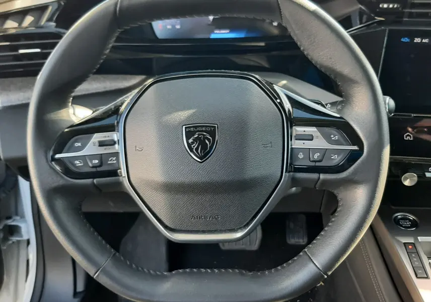 Vue rapprochée du volant noir de la Peugeot 308 2024, avec commandes intégrées et tableau de bord numérique en arrière-plan.