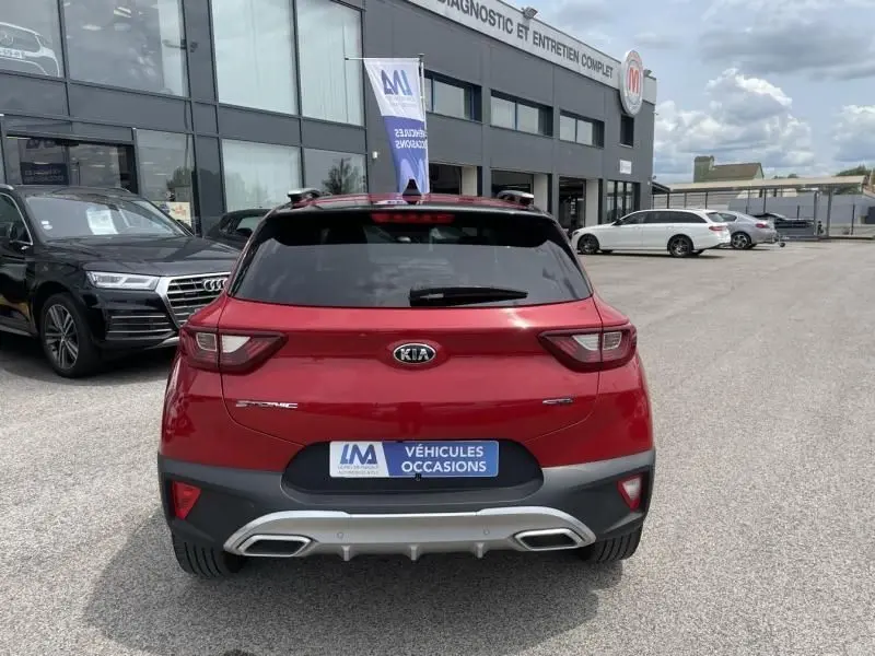 Vue arrière d'un SUV Kia rouge métallisé garé devant un garage de véhicules d'occasion par temps nuageux.