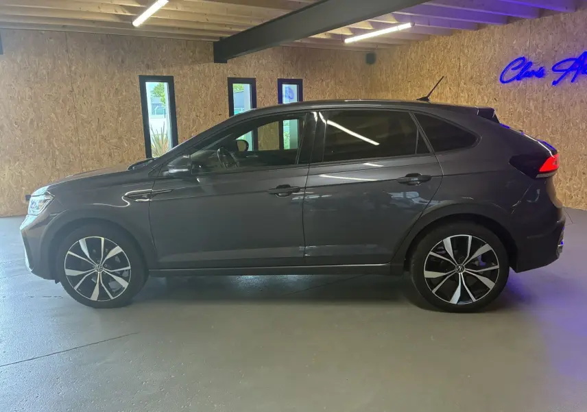 Profil droit d'un Volkswagen Taigo gris 2023 finition R-Line avec jantes alliage et vitres teintées, en intérieur showroom.