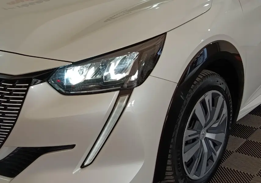 Gros plan sur l'avant droit blanc d'une Peugeot 208 électrique avec phares LED allumés et jante stylisée.