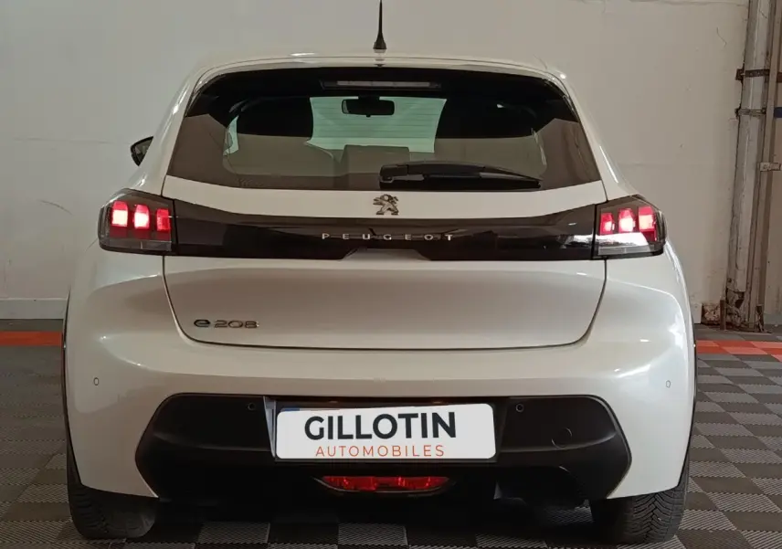 Vue arrière d'une Peugeot 208 électrique blanche avec feux arrière LED allumés dans un garage.