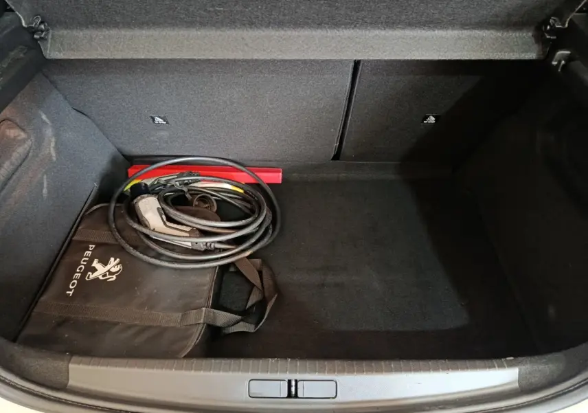 Coffre ouvert d’une Peugeot 208 électrique blanche, avec câble de recharge et sac de rangement Peugeot noir.