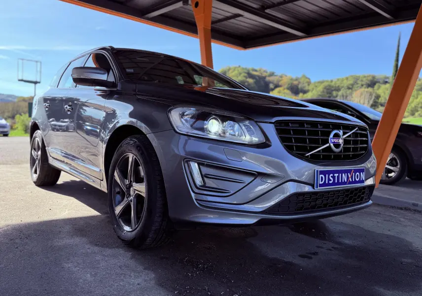 Volvo XC60 gris 3/4 avant droit sous abri, avec phares allumés et calandre noire distinctive.