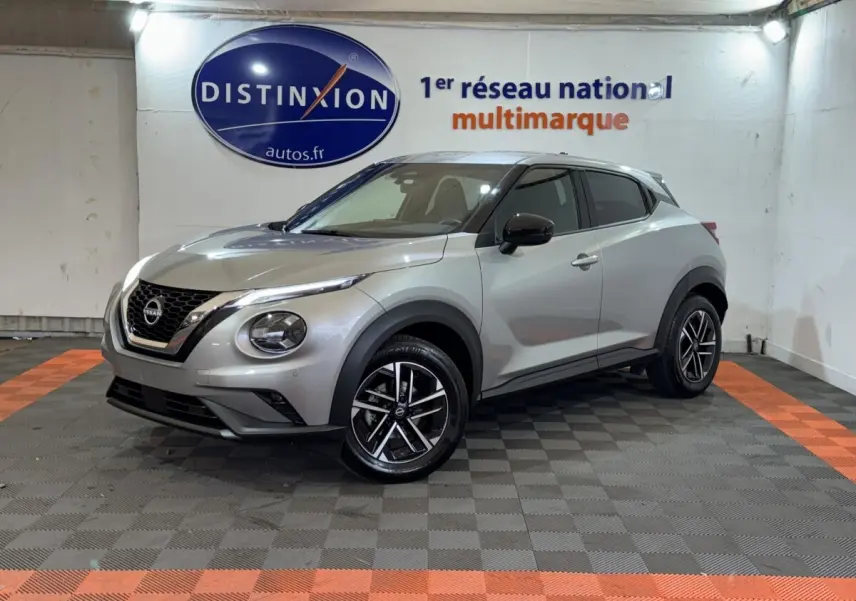 Nissan Juke gris clair vu en 3/4 avant droit, avec jantes alliage noires et calandre V-Motion chromée.