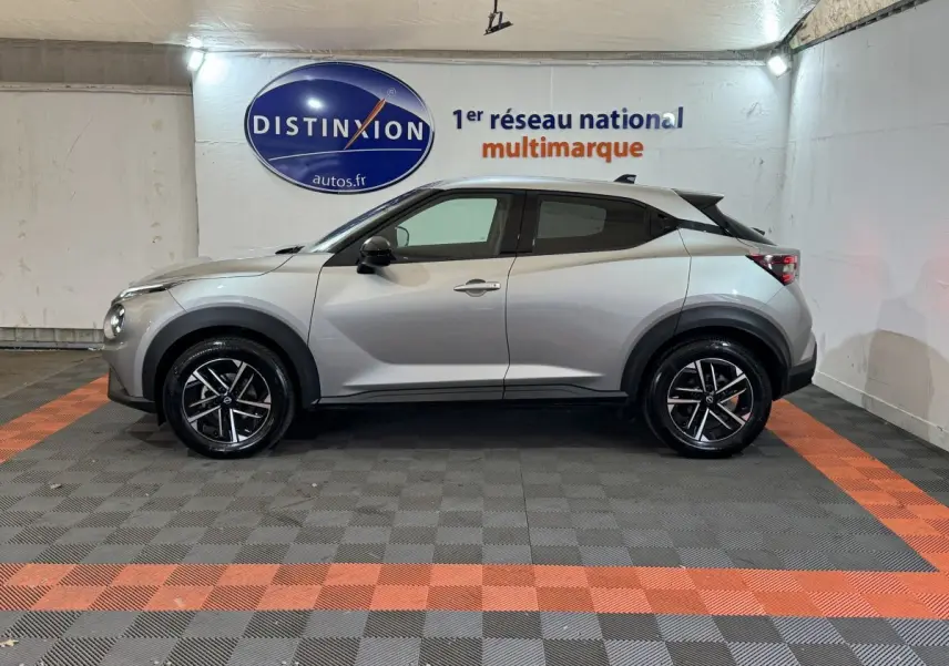 Vue de profil côté gauche d'un Nissan Juke gris clair 2025 avec jantes alliage et toit noir dans un showroom.
