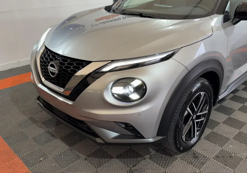 Vue 3/4 avant droit du Nissan Juke gris clair avec phares LED allumés et calandre en V chromée.