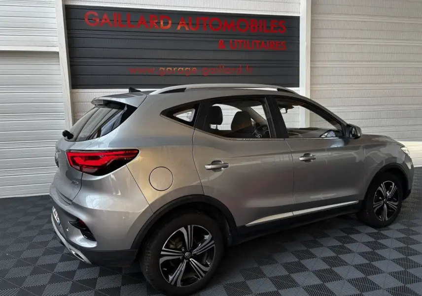 Vue 3/4 arrière côté droit d'un MG ZS gris clair 2022 avec jantes alliage et barres de toit chromées.