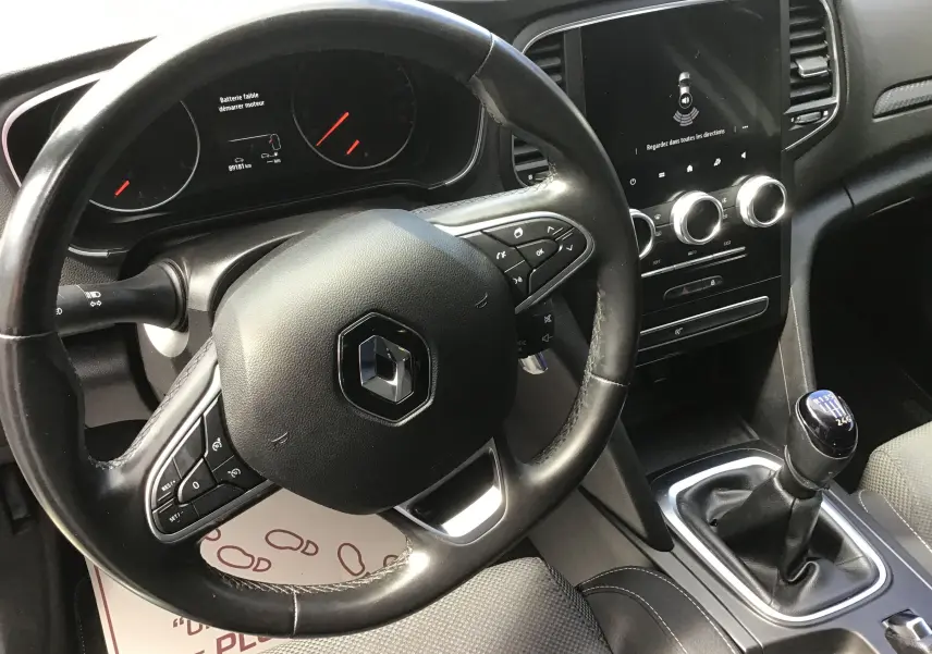 Vue intérieure centrée sur le volant noir et la console centrale avec levier de vitesse manuel d'une Renault Mégane IV Société blanche.
