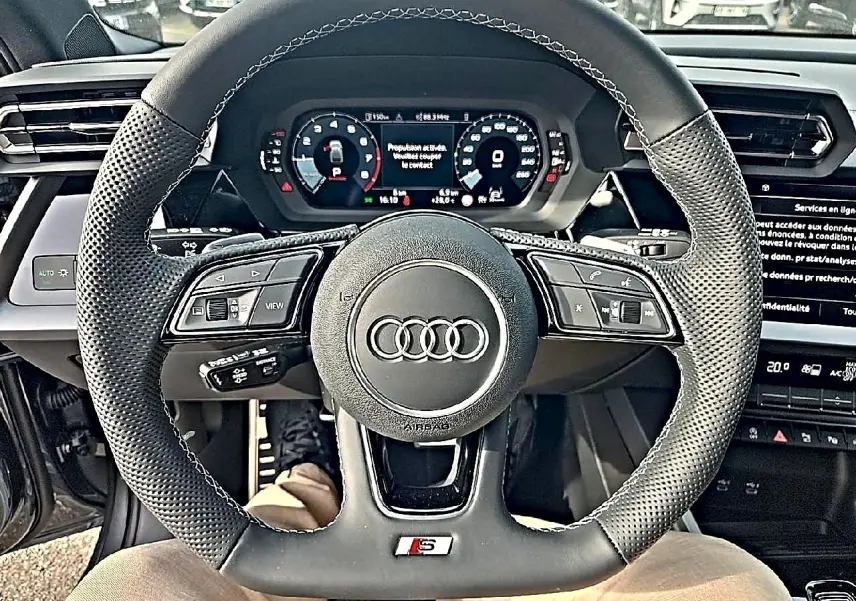 Vue intérieure centrée sur le volant sport noir perforé de l'Audi A3 Sportback 1.5 TFSI S line avec tableau de bord digital.