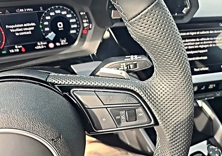 Gros plan sur le volant cuir perforé et les commandes multifonctions de l'Audi A3 Sportback gris Daytona, tableau de bord numérique visible.
