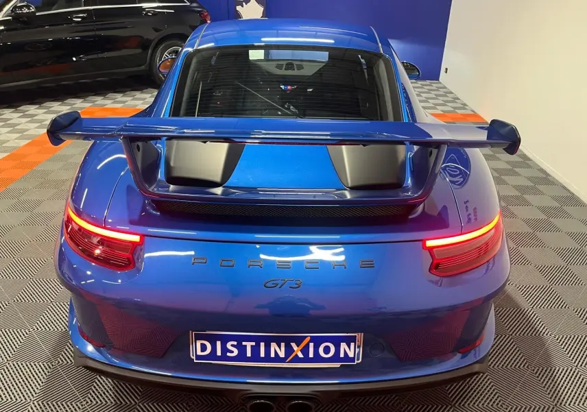 Vue arrière d'une Porsche 911 GT3 bleu saphir avec aileron large et feux arrière allumés en intérieur showroom.