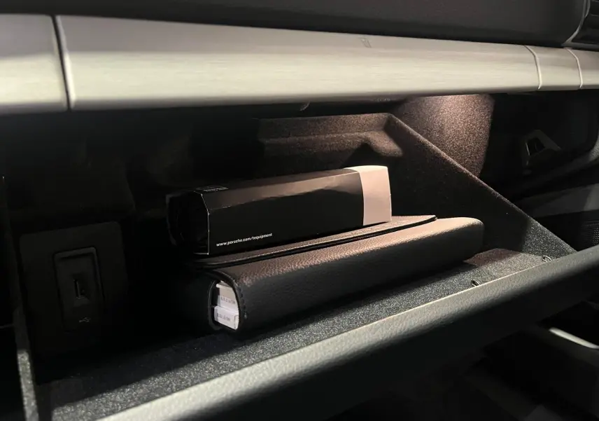 Gant de boîte à gants ouvert avec éclairage intérieur, montrant un manuel Porsche et une boîte noire.
