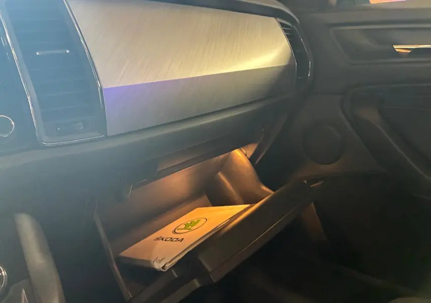 Intérieur du Skoda Kodiaq 2023, vue côté passager sur la boîte à gants ouverte avec manuel Skoda visible, ambiance sombre.