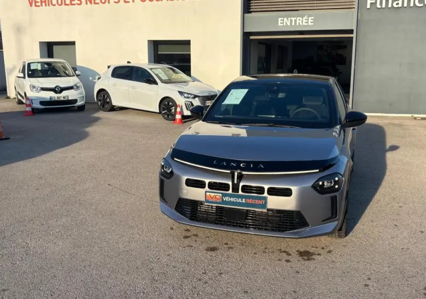 Lancia Ypsilon gris vue de face, stationnée devant un bâtiment avec panneau "Véhicules neufs et occasions".