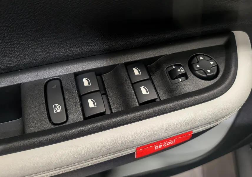 Gros plan sur les commandes de lève-vitres et rétroviseurs électriques du côté gauche, garniture cuir clair avec étiquette rouge "be cool" visible.