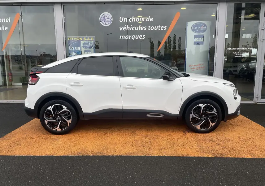 Profil côté gauche d'une Citroën C4 blanche 2022 avec toit noir et jantes alliage distinctives, devant une vitrine commerciale.