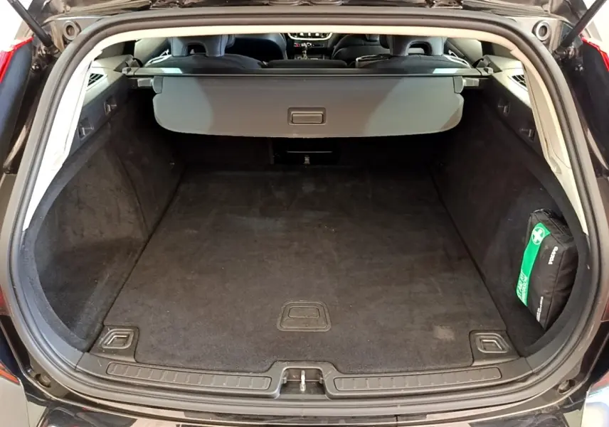 Coffre ouvert vue arrière d’un Volvo V60 noir, avec cache-bagages et trousse de secours visible à droite.
