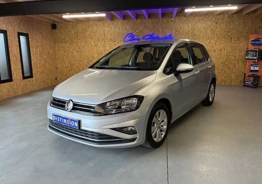 Volkswagen Golf Sportsvan gris vue 3/4 avant droit dans un showroom avec mur en bois et enseigne lumineuse bleue.