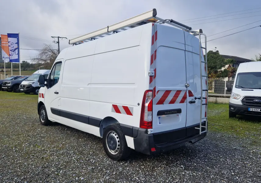 Renault Master blanc en vue 3/4 arrière droit, avec galerie de toit, échelle arrière et bandes réfléchissantes rouges.
