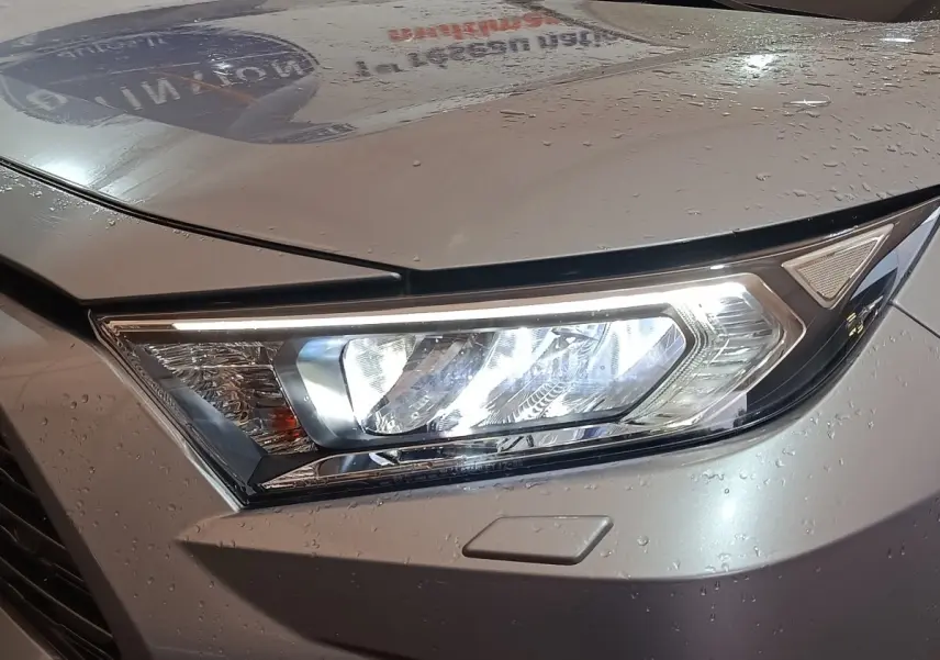 Gros plan sur le phare avant droit allumé d’un Toyota RAV4 hybride gris clair avec gouttes d’eau sur la carrosserie.