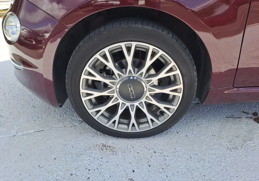 Gros plan sur la roue avant gauche d'une FIAT 500 Opera Bordeaux Métal, mettant en valeur la jante élégante et le phare avant.