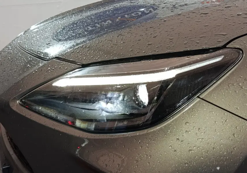 Gros plan sur le phare avant droit allumé d'une Toyota Yaris Cross gris foncé, avec gouttes d'eau sur la carrosserie.
