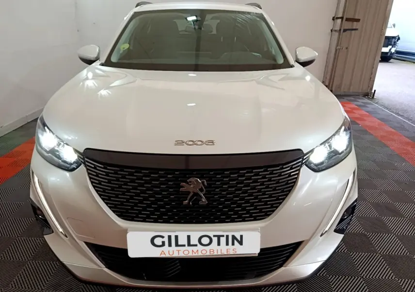 Vue avant d'un Peugeot 2008 blanc avec calandre noire et phares LED allumés dans un garage.