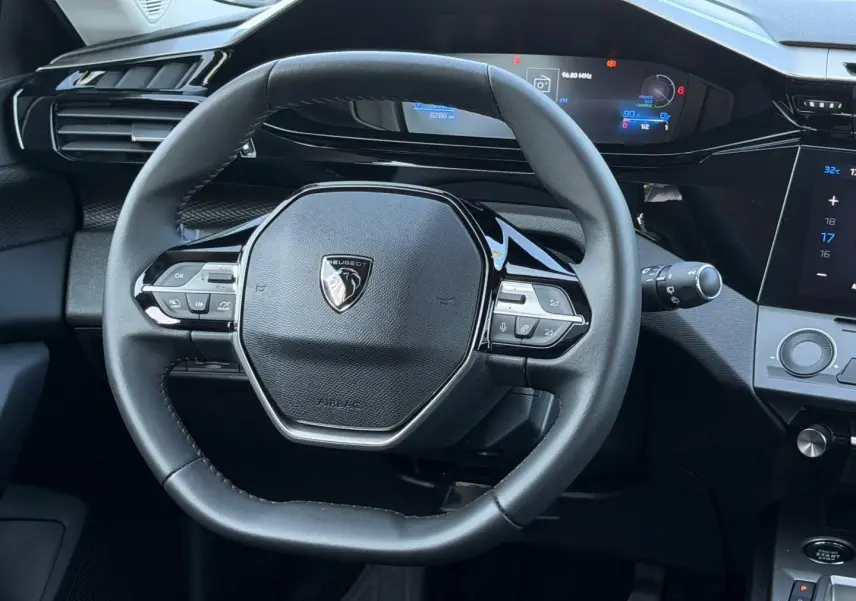 Vue rapprochée du volant multifonction noir de la Peugeot e-308 2025 avec tableau de bord numérique en arrière-plan.