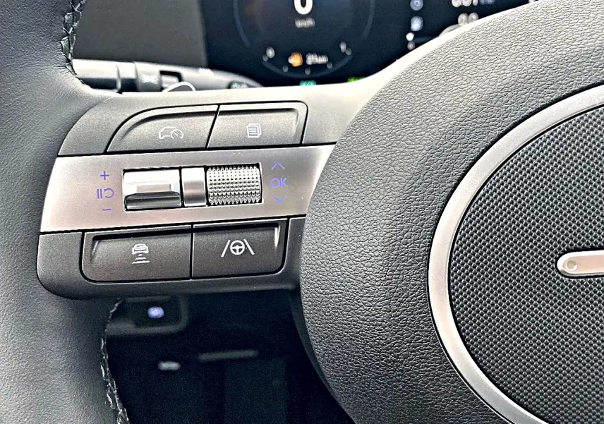 Gros plan sur les commandes à gauche du volant cuir noir du Hyundai Kona gris, avec tableau de bord numérique en arrière-plan.