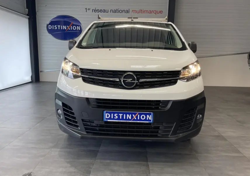 Vue frontale d'un Opel Vivaro blanc avec boucliers noirs et phares allumés dans un showroom.